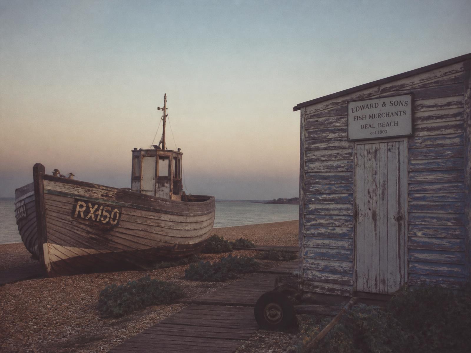 A weathered wooden fishing boat resting on the shingle beside a peeling painted beach hut at dusk